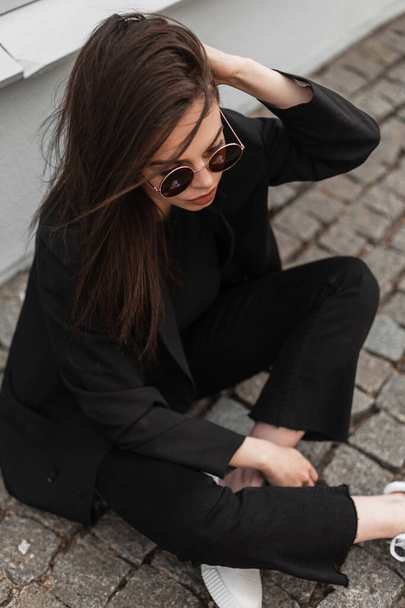 Cool modern young woman in sunglasses in fashionable black casual wear straightens hair near vintage wooden building on street in city. American trendy girl fashion model is resting on stone tile. - Photo, Image