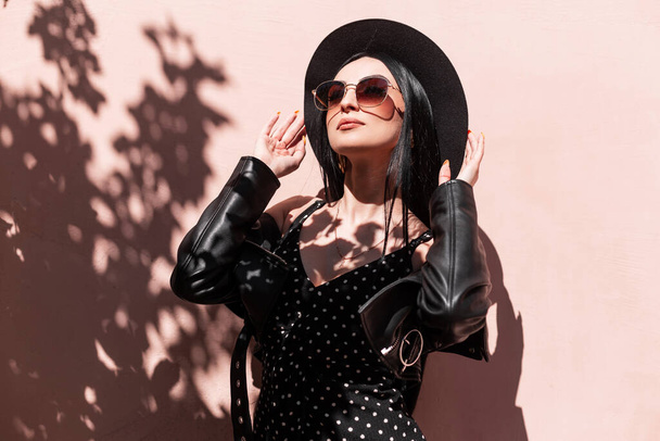 Glamorous young brunette woman in beautiful fashionable black clothes straightens hat outdoors. Luxurious sexy girl in dress in leather jacket in sunglasses posing and enjoys sunlight. Summer style. - Photo, Image