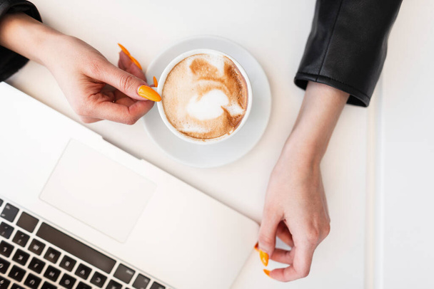 Close-up of a desktop with cup of hot coffee and modern laptop, behind with girl in black stylish clothes sitting. Business woman start coffee break. Remote work. - Photo, Image