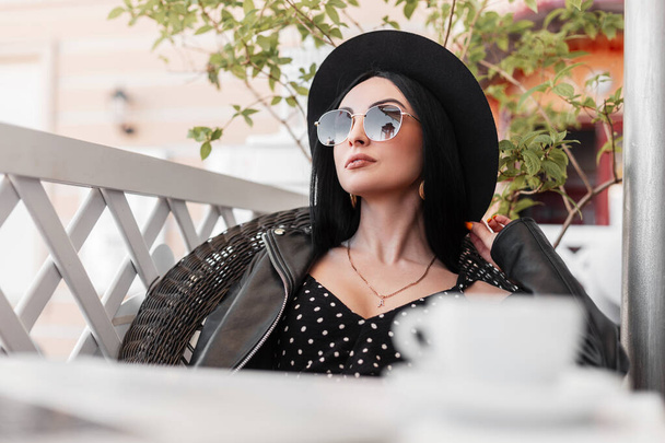Atrakcyjny model moda kobieta w czarnym eleganckim piękne ubrania w okularach przeciwsłonecznych spoczywa na fotelu vintage w letni odkryty kawiarnia w słoneczny dzień. Seksowna dziewczyna zrobiła przerwę na kawę. Dzień dobry.. - Zdjęcie, obraz