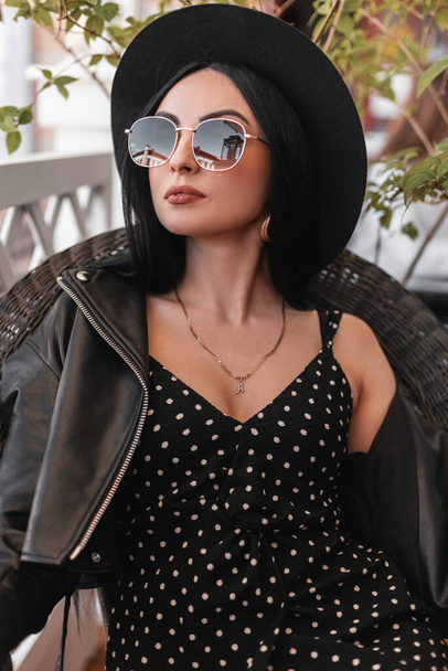 Fresh portrait pretty fashionable woman in black summer dress with hat in sunglasses in leather jacket in cafe. Gorgeous girl in black fashion clothes is resting on vintage chair on summer terrace. - Photo, Image
