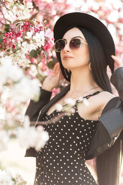 Stylisch schöne junge Model-Frau in trendigen Outfits mit Hut und Sonnenbrille in der Nähe von Blumen am Frühlingstag - Foto, Bild