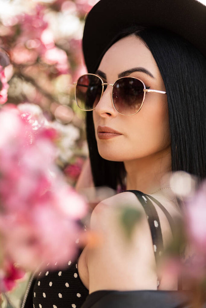 Elegante hermosa mujer joven en ropa de moda con accesorios se encuentra cerca de los árboles con flores - Foto, imagen