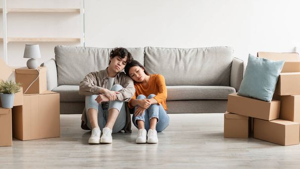 Upset young Asian couple sitting on floor among cardboard boxes, feeling exhausted and sad on moving day, empty space - Фото, зображення