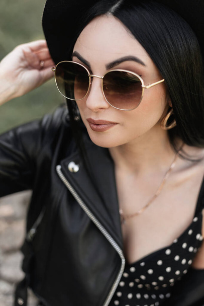 Lovely woman in fashionable sunglasses in elegant hat in black stylish dress in leather casual jacket posing outdoors on sunny spring day. Cute fine girl with beautiful sexy lips enjoy in nature. - Foto, Imagem