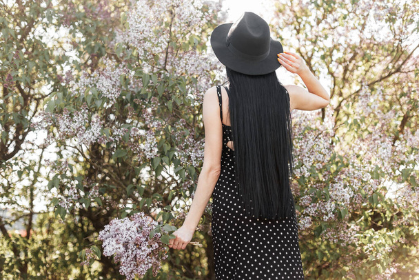 Вид спереду на модну жінку з довгим волоссям у стильній сукні в модному капелюсі з бузковими квітами біля квітучого дерева в парку в сонячний день. Елегантна жінка насолоджується красою природи
. - Фото, зображення