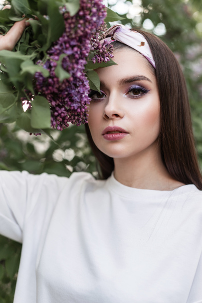 Lovely pretty woman in elegant bandana in white stylish wear stands with amazing color bouquet near green foliage in park. Pretty girl enjoys spring and lilac flowers in garden. Fresh portrait lady. - Foto, Imagen