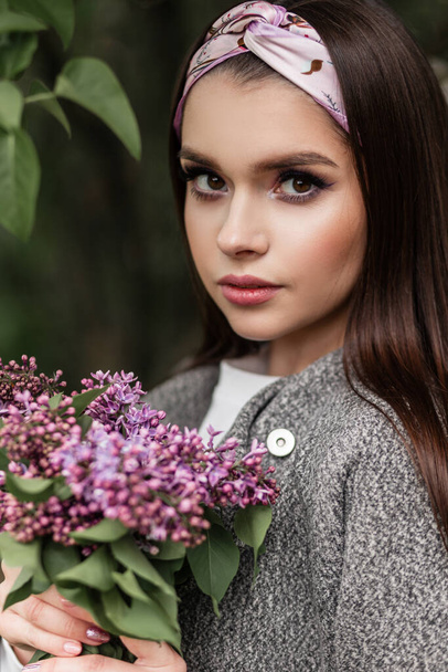 Pretty portrait of woman in bandana in fashionable gray coat with color make-up with bouquet chic purple flowers lilac near green foliage in park. Cute girl with bouquet outdoors - Foto, Imagem
