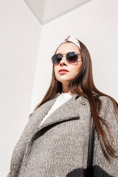 Sunny portrait young beautiful woman in fashionable sunglasses in stylish bandana in gray elegant coat near vintage wall on bright spring day in city. Pretty girl enjoys spring sunlight on street. - Photo, Image