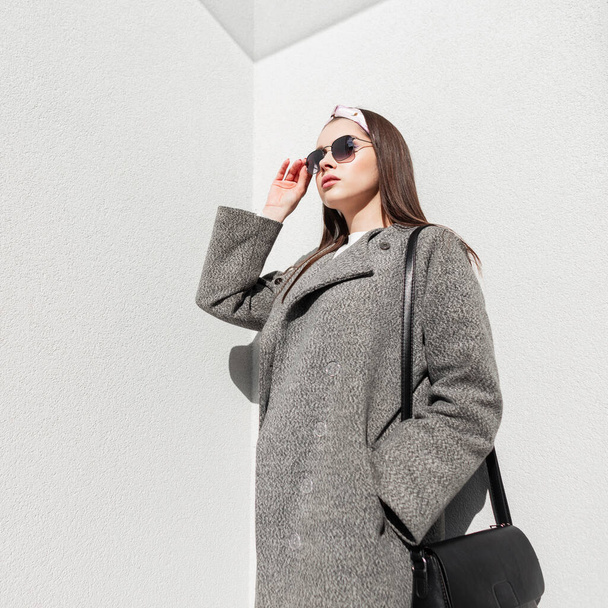 Model glamour woman in gray elegant coat in glamorous bandana in stylish sunglasses relaxes on sun near vintage white wall on street. Beautiful girl in casual outerwear enjoys spring sunlight outdoors - 写真・画像