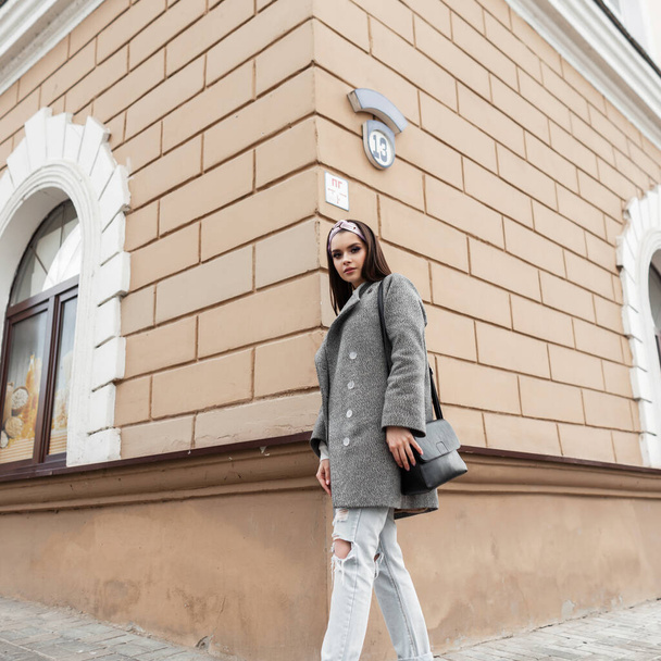Elegant pretty young woman model in trendy bandanna in gray stylish coat with black handbag posing near vintage building outdoors. Beautiful girl in fashionable youth casual spring clothes enjoy walk. - Photo, Image