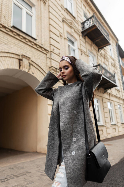 Lovely attractive young woman in glamor bandana in gray beautiful coat with handbag posing near vintage building in city. Stylish gorgeous girl model walks on street. Youth spring fashion womens wear. - Foto, Imagen