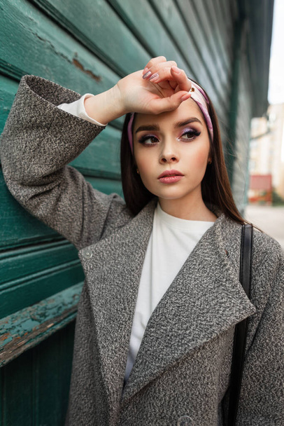 Young gorgeous woman with color make-up in fashionable bandana in gray stylish coat rest near vintage green wooden wall outdoors. Lovely pretty attractive urban girl in fashion casual wear enjoy. - Photo, Image