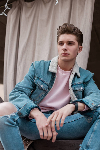 Good-looking young man with trendy hairstyle in fashionable blue youth jeans clothes from new summer collection rest on street outdoors. European stylish guy model in fashion denim casual wear in city - Foto, Imagen