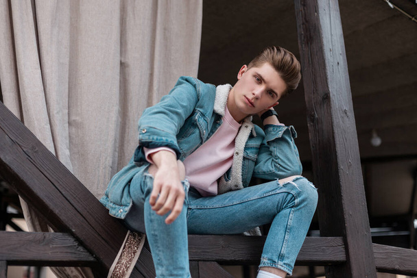 Handsome young man with trendy hairstyle in fashionable blue youth jeans clothes from new summer collection rest on street in city. Attractive serious guy model in fashion denim casual wear outdoors. - Valokuva, kuva