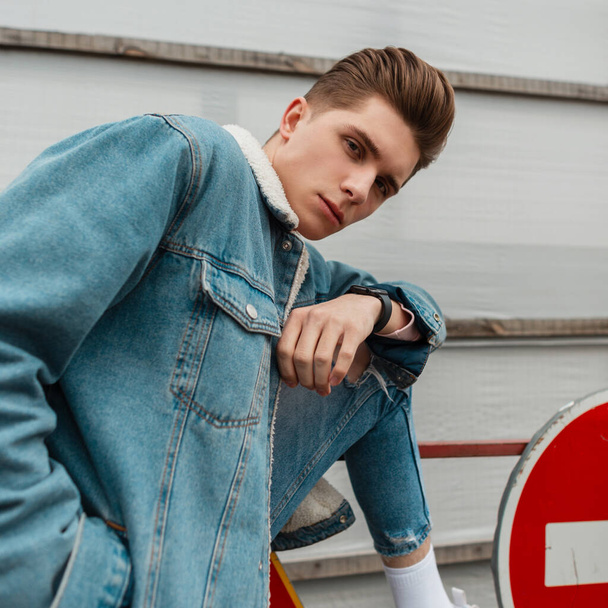 Fashion portrait cool young man with hairstyle in blue youth casual jeans clothes near metal road sign "stop" on street in city. Handsome fashionable guy model in trendy denim outfit outdoors. Summer. - 写真・画像