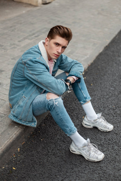 流行のデニムジャケットにちぎれたヴィンテージジーンズの流行の白いスニーカーの現代的な見栄えの若い男は、街の路上で道路の近くのタイルに座っています。クールな都会の男でスタイリッシュな服の残りの部分屋外. - 写真・画像