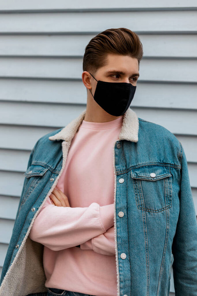 ブラック保護マスク屋外でピンクのスウェットシャツでデニムブルースタイリッシュなジャケットで見栄えの若い男。ファッショナブルな男が街を歩いている。Covid-19ウイルス対策コンセプト。コロナウイルスには - 写真・画像