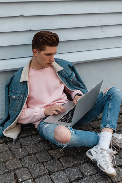 Trendy young man in pink sweatshirt in casual fashionable blue jeans clothes works remotely on laptop sitting on stone tile outdoors. Successful guy modern blogger works on Internet in city.  - Фото, изображение