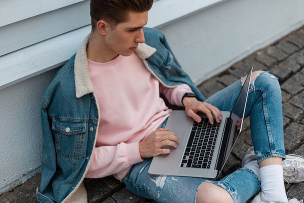Successful young freelancer man in stylish denim casual jeans clothes sits with laptop and works remotely on creative project. Attractive fashion designer is typing on keyboard. Remove work. - Valokuva, kuva