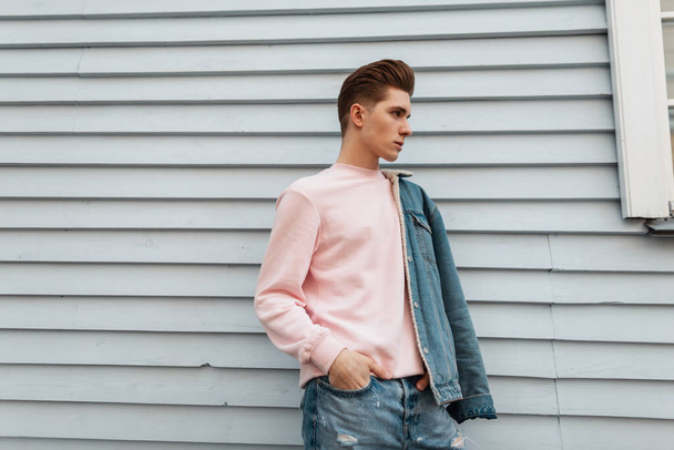 Glamorous dość młody człowiek model w modnej różowej bluzie w drelichu mody niebieskie ubrania młodzieżowe pozowanie w pobliżu białej ściany na zewnątrz. Miejski facet w dżinsach casual strój z nowej kolekcji pozuje na ulicy. - Zdjęcie, obraz
