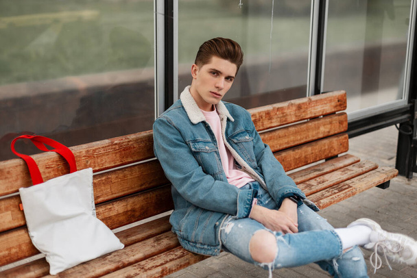 Fashionable young man in casual blue jeans clothes in trendy white sneakers with vintage fabric bag sits on wooden bench at a bus stop in city. Handsome guy in stylish denim youth clothes on street. - Foto, imagen
