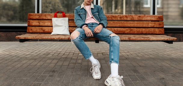 Модний молодий чоловік в юнацькій стильній джинсовій куртці в казуальних вінтажних джинсах з тканинним продавцем сидить на дерев'яній лавці на зупинці громадського будівництва в місті. Гарний хлопець в модному одязі на вулиці
. - Фото, зображення