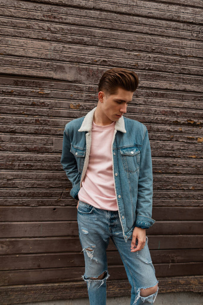 Attractive young man with hairstyle in stylish pink t-shirt in fashionable denim jacket posing near vintage wooden brown wall in street. European handsome guy model in jeans clothes is stand in city. - Photo, Image