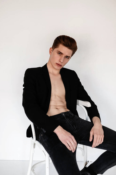 Modieuze man in een zwarte blazer en jeans met een naakte romp zit op een witte stoel in de studio - Foto, afbeelding
