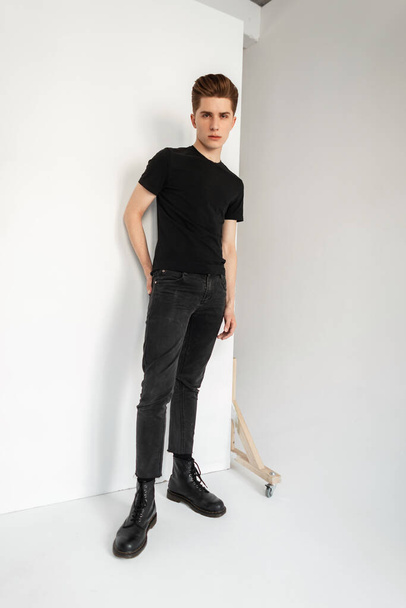 Модна модель стильний молодий чоловік у старовинній чорній футболці в модних сірих молодіжних джинсах, що позують біля білої стіни в кімнаті. Привабливий привабливий хлопець в повсякденному модному одязі стоїть в студії
. - Фото, зображення