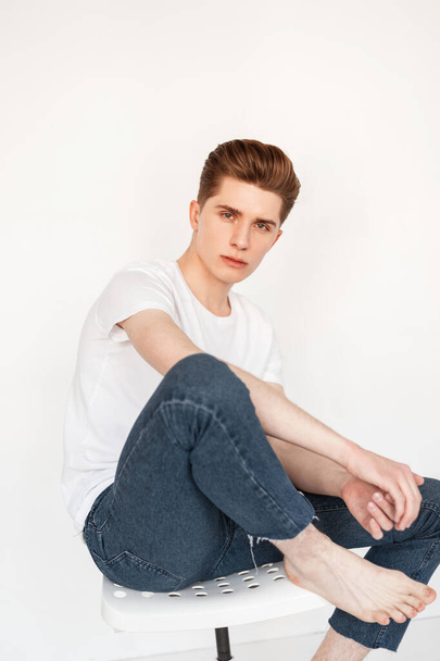 Handsome young man with trendy hairstyle in fashionable white t-shirt in stylish blue jeans sits on chair on white background in studio. Attractive fashion model guy posing near vintage wall in room. - Photo, Image