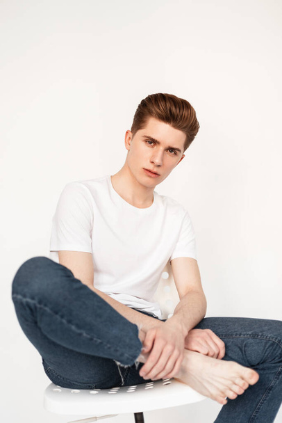 Fashion fresh portrait young man with clean skin with serious face with hairstyle in stylish white t-shirt in blue jeans on white background indoors. Handsome guy is poses on chair near vintage wall. - 写真・画像