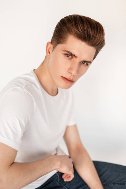 Portret Europese aantrekkelijke jongeman met stijlvol kapsel met schone huid in modieuze klassieke witte t-shirt in blauwe vintage jeans in de buurt van vintage muur in studio. Knappe vent rust op stoel binnen - Foto, afbeelding
