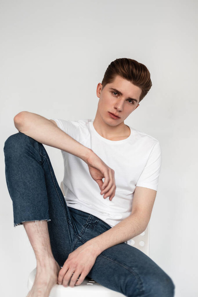 Fashionable serious young man in blue vintage jeans in stylish white t-shirt sits on chair on white chair in room. Studio portrait handsome guy in trendy clothes. New fashion collection casual wear. - Fotografie, Obrázek