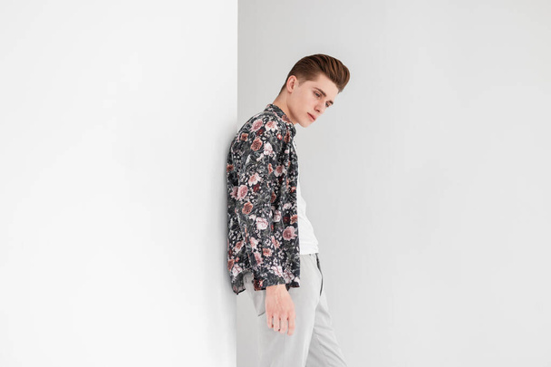 Elegancki młody mężczyzna z modną fryzurą w białej koszulce w kwiatowej koszuli vintage w stylowych spodniach spoczywa w pobliżu białej ściany w studio. Atrakcyjny facet model w odzieży młodzieżowej w pomieszczeniach. - Zdjęcie, obraz