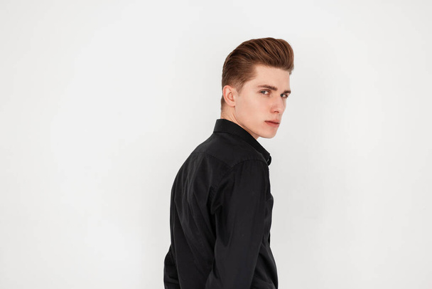 Portrait pretty serious young man with confident face with trendy hairstyle in fashionable elegant black shirt near vintage white wall indoors. Attractive modern fashion model guy in room.Stylish wear - Foto, Imagem