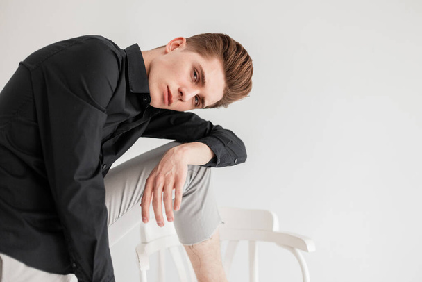Modemodel gut aussehender junger Mann mit trendiger Frisur im modischen schwarzen Hemd in stylischer Hose steht neben Stuhl auf weißem Hintergrund im Atelier. Schöne elegante Kerl posiert in der Nähe Wand drinnen. - Foto, Bild