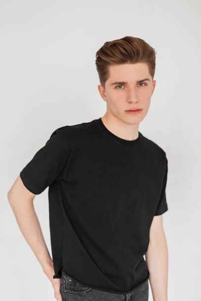 Portrait attractive young man with stylish hairstyle with clean skin in fashionable classic black t-shirt near vintage wall in studio. Handsome guy in trendy wear poses indoors. Casual youth clothing. - Fotó, kép