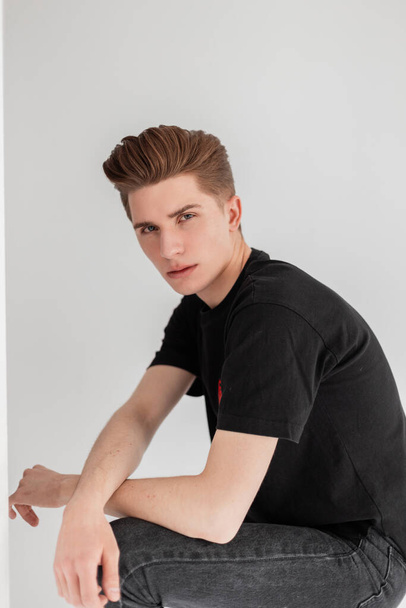 Young serious fashionable man with stylish hairstyle in trendy youth casual black t-shirt from new summer collection stands near vintage wall in studio.Good-looking vogue guy in vintage wear indoors. - Foto, Imagen