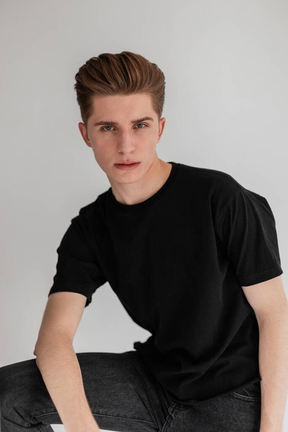 Portrait stylish young man with serious face with trendy hairstyle with clean skin in fashionable black t-shirt on white background in studio. Handsome guy in black summer clothes straightens hair. - Foto, Bild
