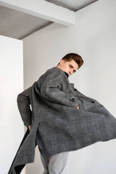 Young stylish man fashion model in luxurious checkered gray trendy coat in vintage pants posing on white wall indoors. Elegant handsome guy in fashionable outerwear in studio. Casual spring menswear. - Foto, Imagem