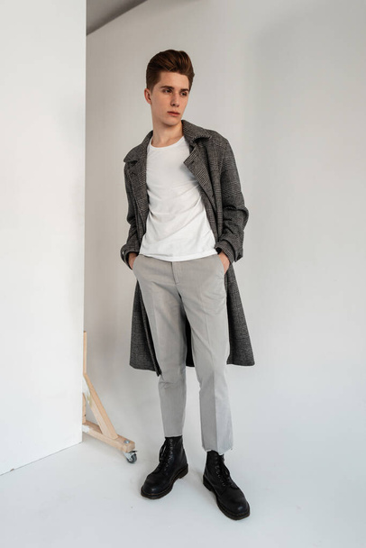流行のコートで白いTシャツの若いファッショナブルな男は、ファッショナブルな革の黒のブーツでエレガントなパンツでコートをプレイスタジオで壁の近くに立っています。 - 写真・画像