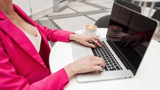 Профессиональная успешная деловая женщина в модной розовой одежде сидит в ярком современном офисе в рабочее время и работает за ноутбуком. Молодая девушка-фрилансер взяла перерыв на кофе. Творческое рабочее место. - Фото, изображение