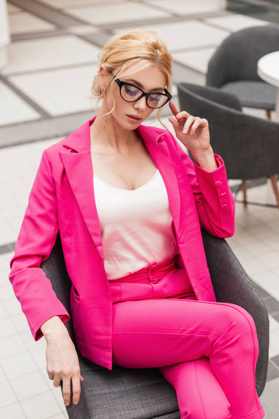 Glamorous όμορφη νεαρή επαγγελματίας γυναίκα σε κλασικό κομψό ροζ κοστούμι με μοντέρνο χτένισμα ισιώνει τα γυαλιά, ενώ κάθεται στην καρέκλα στο γραφείο. Sexy επιχειρηματικό μοντέλο κορίτσι στη συνάντηση στο καφέ. - Φωτογραφία, εικόνα