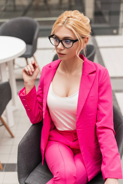 Die schöne junge, stylische Blondine mit modischer Brille in schickem rosa Anzug ruht sich drinnen aus. Modernes Profi-Model in stylischem Outfit sitzt auf einem Stuhl im Café. Sexy Dame. - Foto, Bild