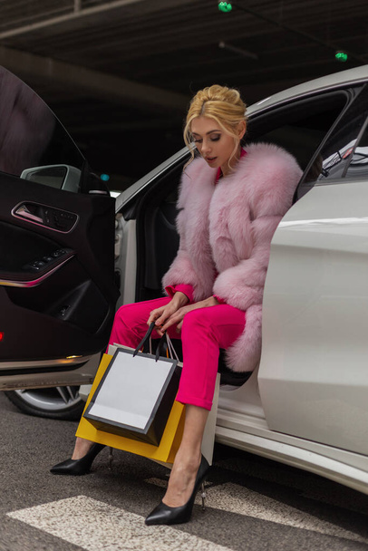 Довольно стильная молодая блондинка в шикарном стильном розовом шубе в штанах позирует с бумажными пакетами в современном автомобиле. Модная привлекательная сексуальная модель девушки останавливается в машине на парковке. Элегантная леди. - Фото, изображение