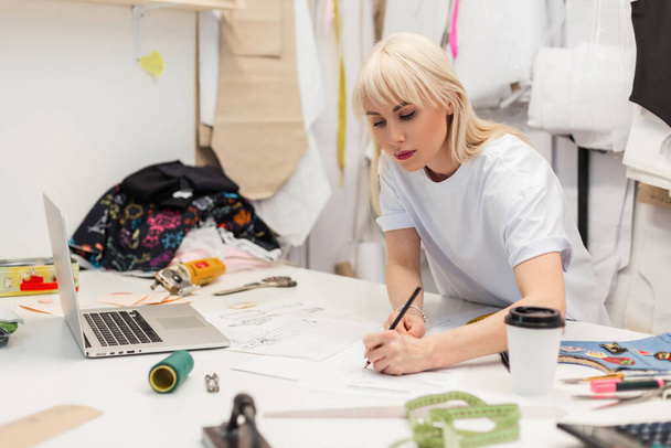Αρκετά σοβαρό ράφτη σε λευκό t-shirt που εργάζονται κοντά στο ξύλινο τραπέζι με ένα φορητό υπολογιστή, καφέ, ψαλίδι, νήμα, εκατοστό και άλλα εργαλεία ραπτικής. Δημιουργικός σχεδιαστής μόδας σχεδιάζει ένα σκίτσο - Φωτογραφία, εικόνα