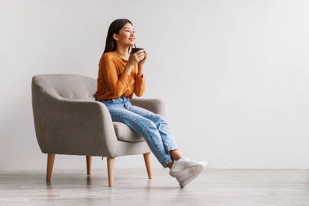 Красивая молодая азиатка пьет кофе или чай, отдыхает в кресле у белой стены студии - Фото, изображение