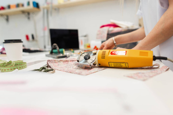 Młody kreatywny krawiec z elektrycznym nożykiem tnie tkaninę w studio. Obszar roboczy projektanta odzieży z laptopem, nitkami, tkaniną, nożyczkami i centymetrem. - Zdjęcie, obraz