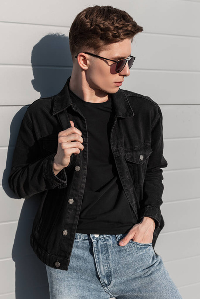 Модний американський молодий гіпстер у вінтажних сонцезахисних окулярах в юному стилі, одягнений в джинсовий одяг з рюкзаком, відпочиває біля будівлі в сонячний весняний день. Привабливий хлопець моделює на вулиці. - Фото, зображення